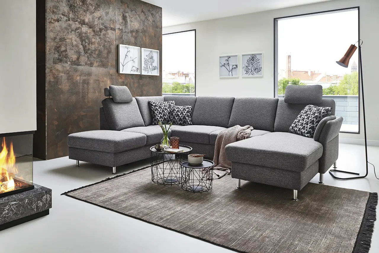 Große graue Wohnlandschaft mit Ottomane im modernen Wohnzimmer mit Kamin, Teppich, Couchtischen und großen Fenstern als Beispiel für stilvolle Wohnzimmergestaltung mit komfortablem Ecksofa